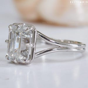 May include: A silver-toned ring featuring a large, rectangular emerald-cut clear gemstone held by eight prongs. The band has a split-shank design that connects to the setting. The text ELITEMOISSY HANDMADE JEWELLERY is visible in the corner.