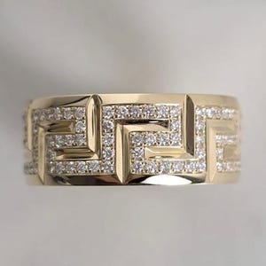 May include: A gold-colored ring with a geometric pattern. The ring features a repeating Greek key design, encrusted with small, sparkling gemstones. The ring is set against a soft, neutral background.