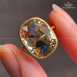 May include: A gold ring featuring a large, cushion-cut gemstone with multiple facets. The gemstone has a mix of colors, including yellow, brown, and blue. The ring's band is a simple, thin gold band.