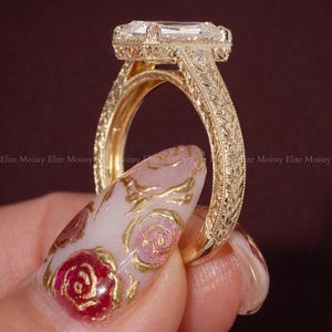 Op de afbeelding: Een close-up van een goudkleurige ring met een smaragdgeslepen edelsteen. De ring heeft ingewikkelde gegraveerde details op de band en zetting. De achtergrond is donkerrood. De merknaam "ELITEMOISSY HANDMADE JEWELLERY" is zichtbaar.