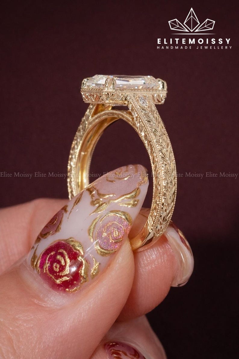 Op de afbeelding: Een close-up van een goudkleurige ring met een smaragdgeslepen edelsteen. De ring heeft ingewikkelde gegraveerde details op de band en zetting. De achtergrond is donkerrood. De merknaam "ELITEMOISSY HANDMADE JEWELLERY" is zichtbaar.