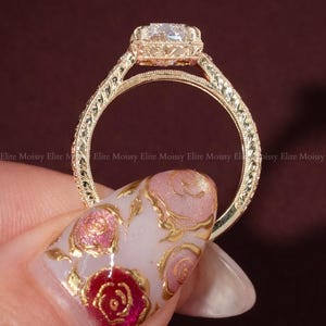 Op de afbeelding: Een goudkleurige ring met een vierkant geslepen edelsteen. De ring heeft ingewikkelde filigraan details op de band en rond de edelsteen. De merknaam "ELITEMOISSY HANDMADE JEWELLERY" is zichtbaar.