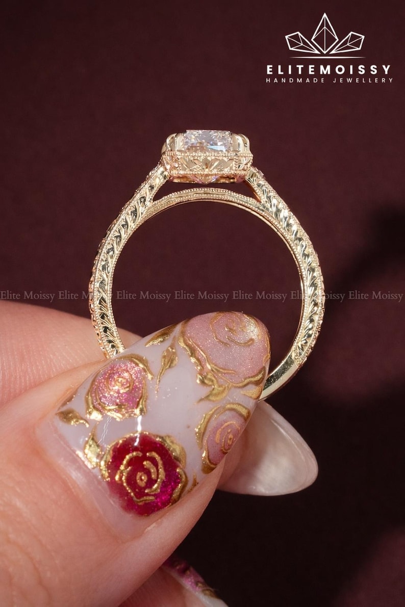 Op de afbeelding: Een goudkleurige ring met een vierkant geslepen edelsteen. De ring heeft ingewikkelde filigraan details op de band en rond de edelsteen. De merknaam "ELITEMOISSY HANDMADE JEWELLERY" is zichtbaar.