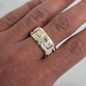 May include: A gold-colored ring with a geometric design and small, sparkling stones. The ring features a repeating pattern of squares and lines, and is worn on a finger.