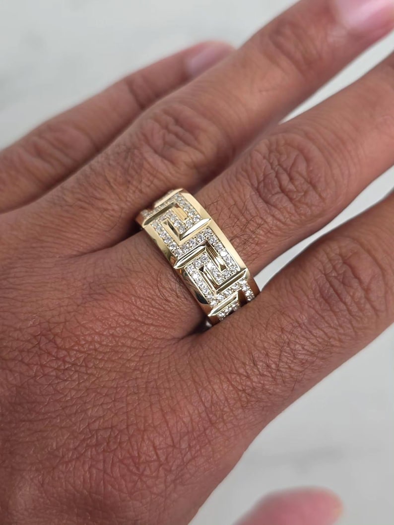 May include: A gold-colored ring with a geometric design and small, sparkling stones. The ring features a repeating pattern of squares and lines, and is worn on a finger.