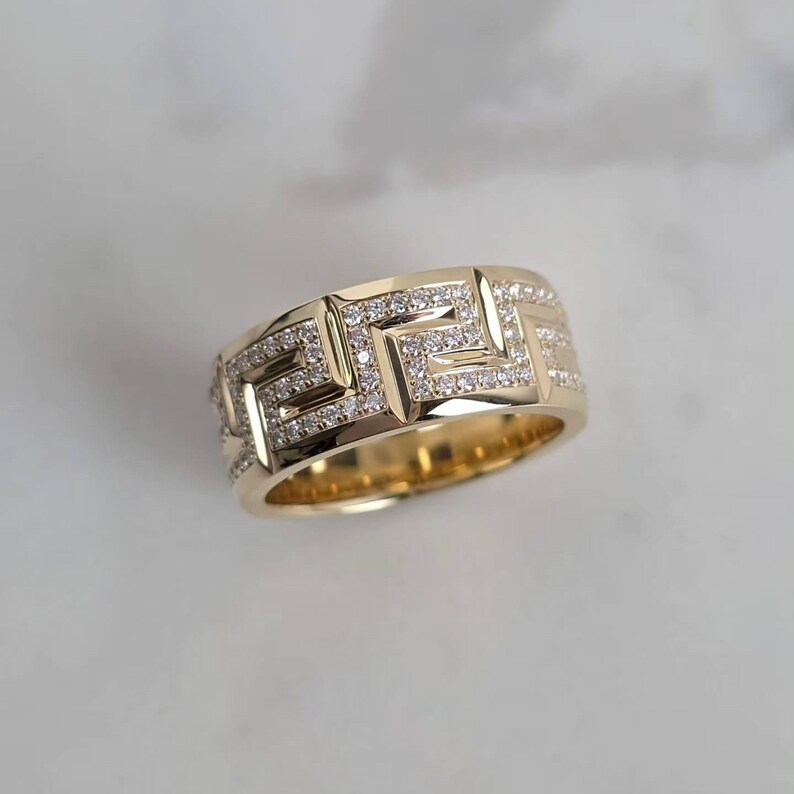 May include: A gold-colored ring with a geometric design. The ring features a repeating pattern of squares and lines, encrusted with small, sparkling clear stones. The ring is set against a white background.