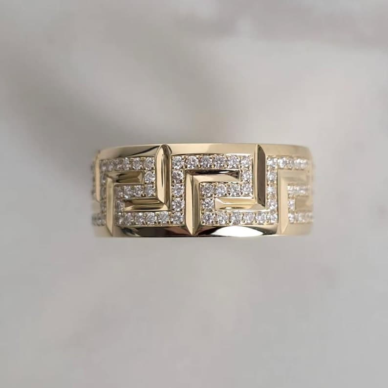 May include: A gold-colored ring with a geometric pattern. The ring features a repeating Greek key design, encrusted with small, sparkling gemstones. The ring is set against a soft, neutral background.