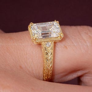 Op de afbeelding: Een goudkleurige ring met een grote, rechthoekige, heldere edelsteen. De ring heeft een sierlijke, gegraveerde band en zetting. De ring is handgemaakt en draagt de merknaam "ELITEMOISSY HANDMADE JEWELLERY".