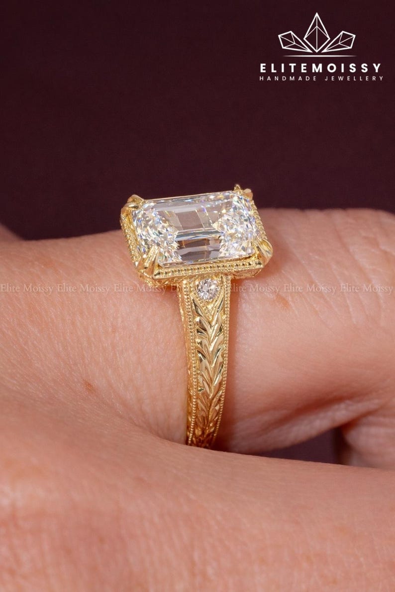 Op de afbeelding: Een goudkleurige ring met een grote, rechthoekige, heldere edelsteen. De ring heeft een sierlijke, gegraveerde band en zetting. De ring is handgemaakt en draagt de merknaam "ELITEMOISSY HANDMADE JEWELLERY".
