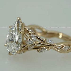 May include: A gold ring featuring a pear-shaped, clear gemstone as the primary stone. The ring's band and setting are adorned with smaller, marquise-cut stones and a delicate, leaf-like design. The ring is set against a soft, neutral background.