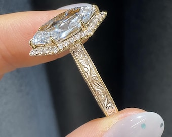 May include: A gold ring featuring a large marquise-cut center stone surrounded by a halo of small round diamonds. The band is crafted from yellow gold and displays intricate, engraved scrollwork patterns along the sides of the shank.
