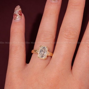 Op de afbeelding: Een gouden ring met een rechthoekige, heldere edelsteen wordt op een hand getoond. De ring heeft een gedetailleerde band en een halo van kleinere stenen rond de hoofdsteen. De nagels zijn beschilderd met een bloemenontwerp. De tekst "ELITEMOISSY HANDMADE JEWELLERY" is zichtbaar.