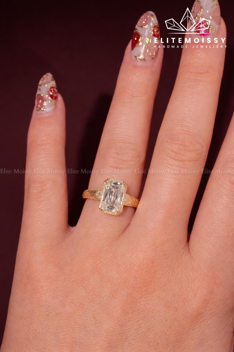 Op de afbeelding: Een gouden ring met een rechthoekige, heldere edelsteen wordt op een hand getoond. De ring heeft een gedetailleerde band en een halo van kleinere stenen rond de hoofdsteen. De nagels zijn beschilderd met een bloemenontwerp. De tekst "ELITEMOISSY HANDMADE JEWELLERY" is zichtbaar.