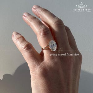 Puede incluir: Un anillo de oro con un gran diamante de talla ovalada. El anillo tiene un diseño de caña dividida. El texto "pretty unreal front view" es visible. El anillo está en una mano.