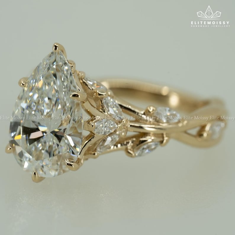 May include: A gold engagement ring featuring a large, pear-shaped diamond as the centerpiece. The band has a twisted design with smaller accent diamonds and leaf-like details. The ring is set against a neutral background.