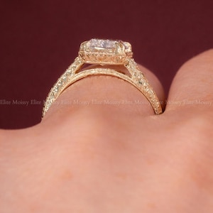 Op de afbeelding: Een goudkleurige ring met een grote, vierkant geslepen edelsteen. De ring heeft ingewikkelde filigraan details op de band en rond de edelsteen. De tekst "ELITEMOISSY HANDMADE JEWELLERY" is zichtbaar.