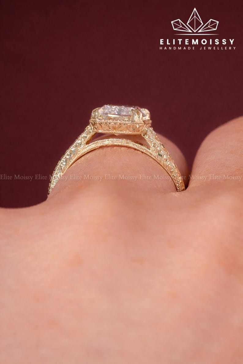 Op de afbeelding: Een goudkleurige ring met een grote, vierkant geslepen edelsteen. De ring heeft ingewikkelde filigraan details op de band en rond de edelsteen. De tekst "ELITEMOISSY HANDMADE JEWELLERY" is zichtbaar.
