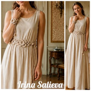 Op de afbeelding: Een mouwloze beige maxi-jurk met een ronde halslijn en een bijpassende gehaakte riem met bloemmotieven en kwastjes. De jurk wordt gecombineerd met een bijpassende gehaakte polsband met bloemmotief voor een samenhangend geheel.