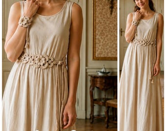 May include: A sleeveless beige maxi dress featuring a round neckline and a matching crocheted belt with floral motifs and dangling tassels. The dress is paired with a coordinating crocheted floral wrist cuff, creating a cohesive look.