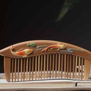 May include: Wooden comb with carved koi fish and cloud designs. The comb features a curved top and fine teeth. The koi fish are painted in orange, yellow, and white, with green and blue accents. A decorative hair accessory.