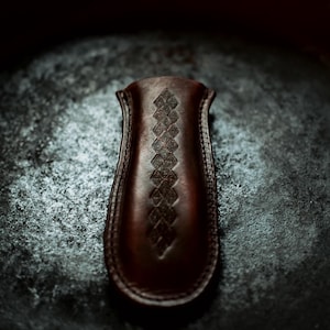May include: A dark brown leather sheath with a decorative diamond pattern. The sheath is curved and has a smooth, polished finish. The background is a dark, textured surface.