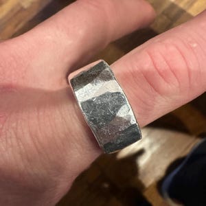 May include: A silver ring with a textured, hammered finish. The ring is wide and rectangular, with a slightly uneven surface. The metal has a bright, reflective quality, and the ring is worn on a finger.