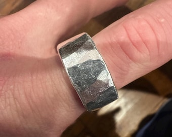 Könnte beinhalten: Ein silberner Ring mit strukturierter, gehammerter Oberfläche. Der Ring ist breit und rechteckig, mit einer leicht unebenen Oberfläche. Das Metall hat eine helle, reflektierende Qualität, und der Ring wird an einem Finger getragen.