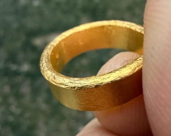 May include: Close-up of a textured gold ring. The ring has a thick band with a rough, hammered appearance. The gold color is bright and shiny, reflecting light. The ring is held between fingers against a blurred green background.