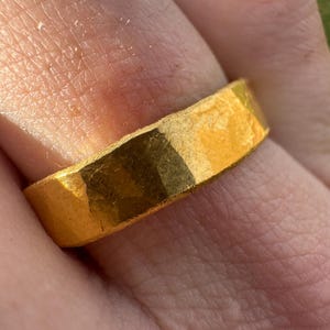 May include: A gold-colored ring with a textured surface. The ring is a band style, with a flat top and sides. The ring is worn on a finger, and the skin around the ring is visible.