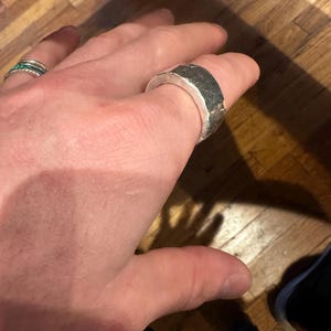 May include: A silver ring with a textured, dark gray band. The ring is thick and has a geometric design. Another ring with small green stones is also visible.