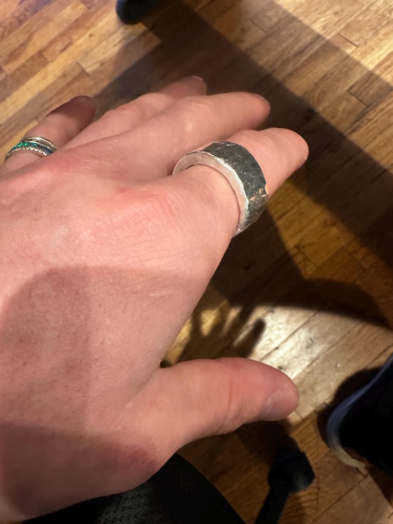 May include: A silver ring with a textured, dark gray band. The ring is thick and has a geometric design. Another ring with small green stones is also visible.