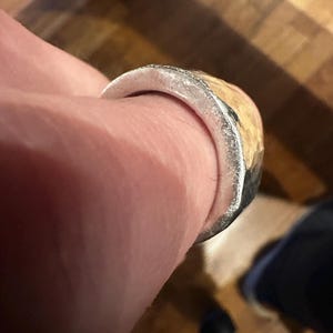 May include: A silver ring with a textured, light-colored band. The ring has a hammered metal appearance and is worn on a finger. The ring's design is simple and rustic.