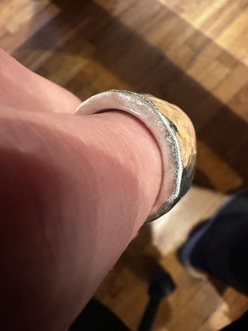 May include: A silver ring with a textured, light-colored band. The ring has a hammered metal appearance and is worn on a finger. The ring's design is simple and rustic.