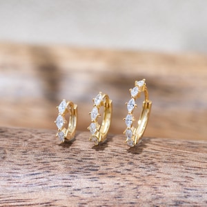 May include: Three gold hoop earrings of varying sizes arranged in a row. Each hoop features a series of clear, marquise-cut stones set along the outer edge. The metal has a polished gold finish, and the hoops are designed with a hinged closure mechanism.