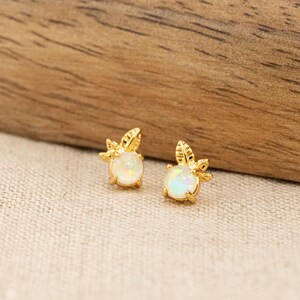May include: A pair of gold stud earrings featuring a round, iridescent white opal stone set in the center. Each earring is accented with a small, textured gold leaf design extending from the top of the circular stone setting.