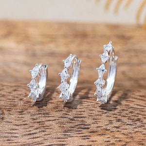 May include: Three silver hoop earrings arranged in a row, each featuring a series of marquise-cut clear stones set along the outer edge. The hoops vary in size, with the smallest on the left and the largest on the right, showcasing a metallic finish.