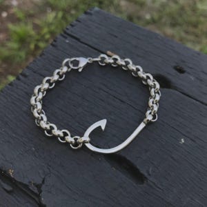 Puede incluir: Una pulsera plateada con un diseño de anzuelo. La pulsera tiene un estilo de cadena con un cierre de mosquetón. El anzuelo tiene una forma curva prominente, añadiendo un toque náutico único a la joya. La pulsera es de metal plateado.