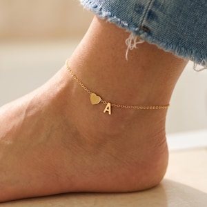 Könnte beinhalten: Ein goldfarbenes Fußkettchen mit einem Herzanhänger und dem Buchstaben "A", der an einer zarten Kette hängt. Das Fußkettchen wird am Knöchel getragen, mit einem Teil einer blauen Jeans im Hintergrund.