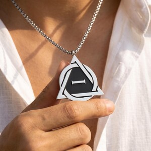 May include: A silver pendant necklace with a triangular design and a black centre, featuring the letter "I". The pendant is suspended from a chain necklace. The necklace is worn against a white shirt.