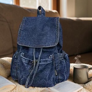 Puede incluir: Una mochila de mezclilla azul con cierre de solapa y cordón. La mochila tiene dos bolsillos laterales y un bolsillo frontal. Una pequeña etiqueta con la palabra "CLADOR" es visible en el lateral. La mochila está sobre una superficie blanca y suave.