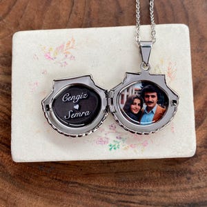 Puede incluir: Collar con colgante en forma de concha plateada, abierto para revelar una fotografía y los nombres "Cengiz Semra" con un corazón. El colgante cuelga de una delicada cadena, sobre una superficie con estampado floral.