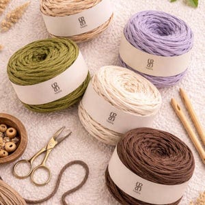 May include: A collection of yarn skeins in various colors, including green, beige, brown, and purple. Each skein is wrapped with a white label. Also included are gold scissors, wooden beads, and knitting needles.