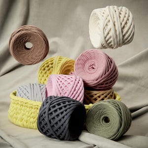 Può includere: Una collezione di gomitoli di filato colorati in varie tonalità, tra cui rosa, giallo, grigio e verde. Alcuni sono disposti in un cestino giallo, mentre altri fluttuano in aria. Il filato sembra essere un materiale spesso e strutturato.