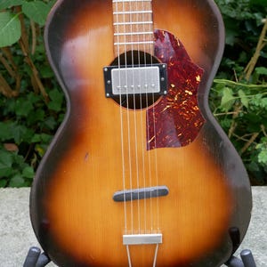 May include: An acoustic-electric guitar with a sunburst finish, featuring a dark brown to light amber gradient. It has a single silver humbucker pickup, a tortoiseshell-patterned pickguard, a wooden bridge, a metal tailpiece, and six steel strings.