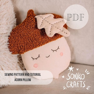 Op de afbeelding: Een naaipatroon en handleiding voor een eikelkussen van Sonko Crafts. De eikel heeft een getextureerde bruine dop, een lichtbeige basis met een gestikt slapend gezichtje en roze wangen, en een klein beige blaadje aan de bovenkant. Tekst PDF.