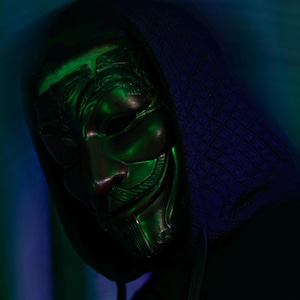 May include: A person wearing a black hooded sweatshirt and a metallic Guy Fawkes mask. The mask has a stylized face with a smile. The background is a blur of green and blue.