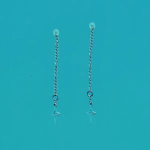Peut inclure: Une paire de boucles d'oreilles à chaîne argentées. Chaque boucle d'oreille est composée d'une fine chaîne suspendue à un petit charme en forme d'étoile à quatre branches. Sur fond turquoise uni.