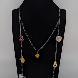 Op de afbeelding: Een zilverkleurige ketting met meerdere kleurrijke emaille bedels, waaronder een geel smileygezicht, rode kersen, een gele citroen, een witte margriet en een groene avocado, met zilverkleurige oordopjes aan de uiteinden van de ketting.