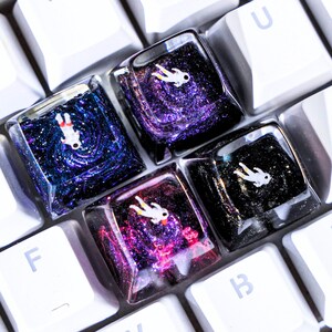 May include: Four square mechanical keyboard keycaps featuring a deep space theme with swirling glittery nebula patterns in shades of blue, purple, and black. Each translucent resin keycap contains a small white astronaut figurine floating inside.