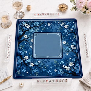 Puede incluir: Un tapete de mahjong cuadrado con fondo azul profundo, ramas oscuras y flores de cerezo blancas. El centro tiene un cuadrado azul sólido con un borde decorativo. Cuatro soportes de acrílico transparente para fichas rodean el tapete.